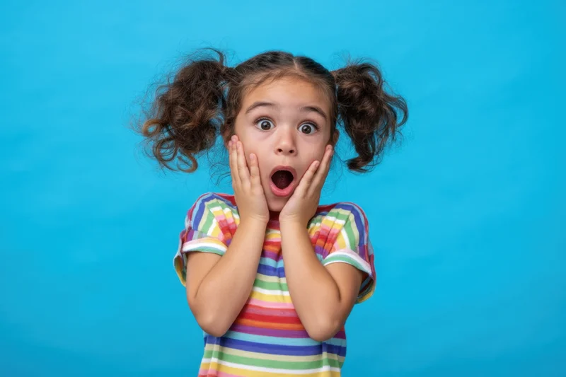 Young girl with pigtails in rainbow-striped shirt showing surprised expression against blue background