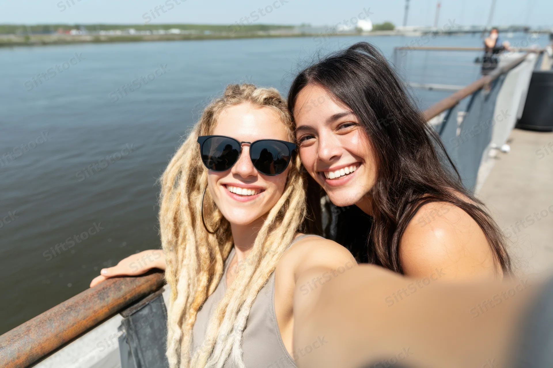 Two women smiling for a selfie by a railing along a waterfront dock