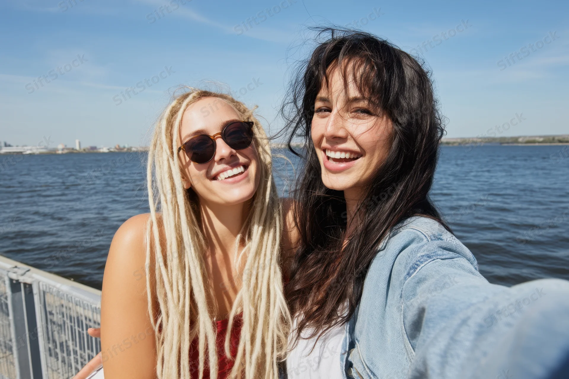 Two women smiling for a selfie by a railing along the waterfront on a sunny day