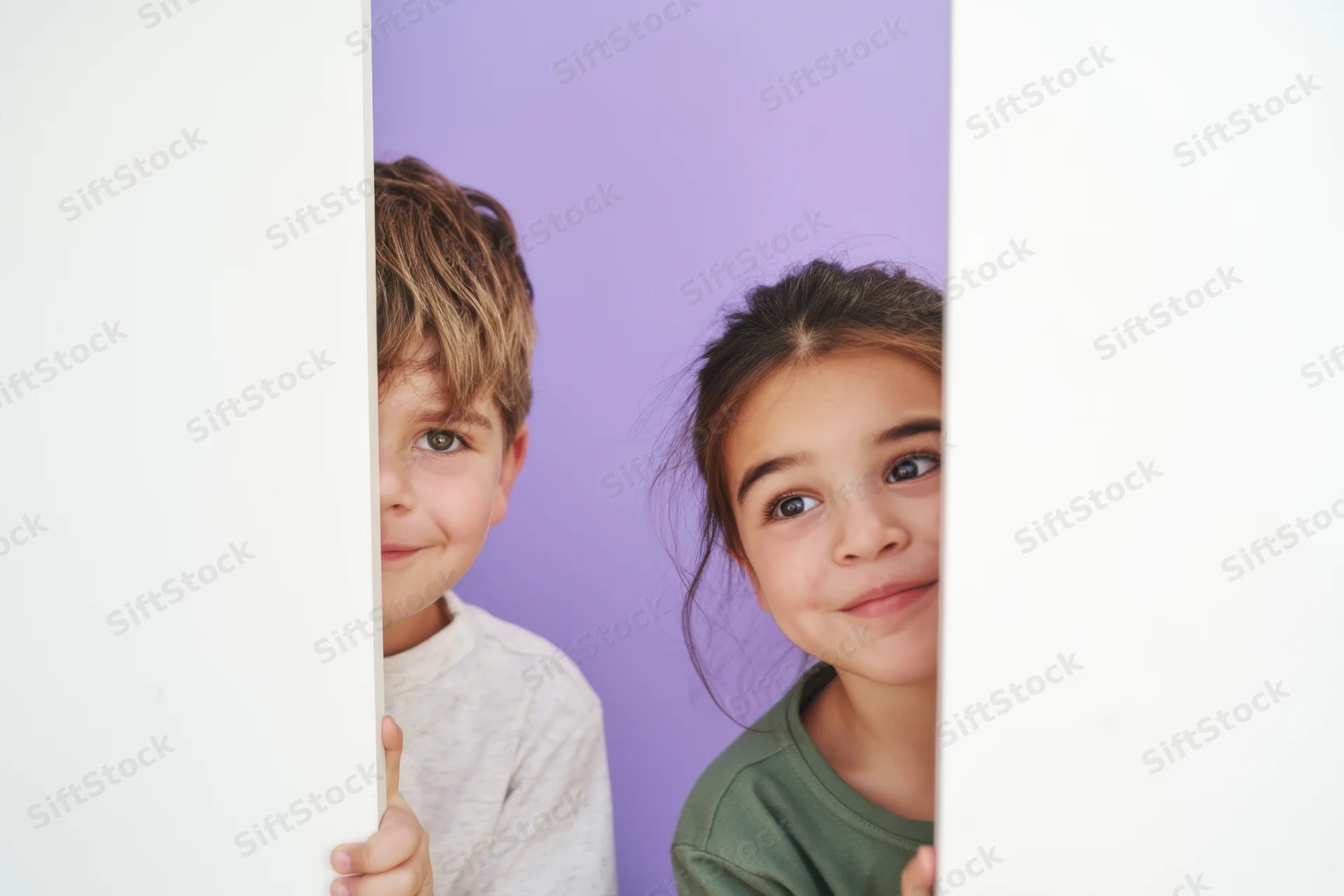 Two young children peeking from behind a tall white panel in a studio setting