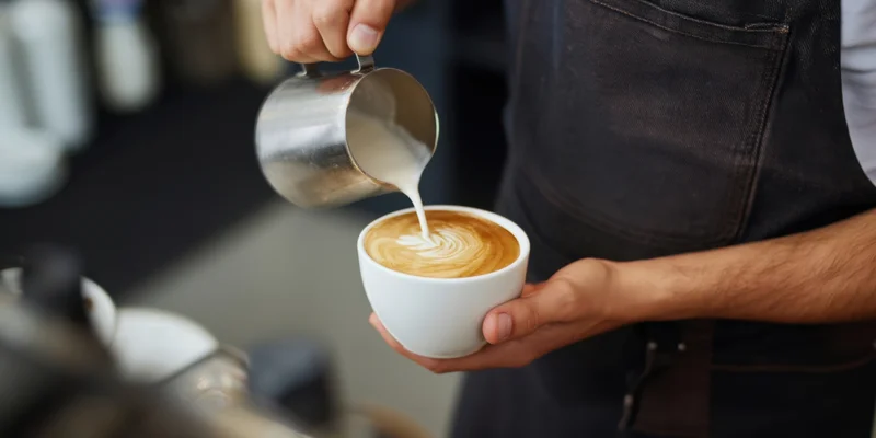 Barista pouring steamed milk into a latte cup to create foam art in a coffee shop