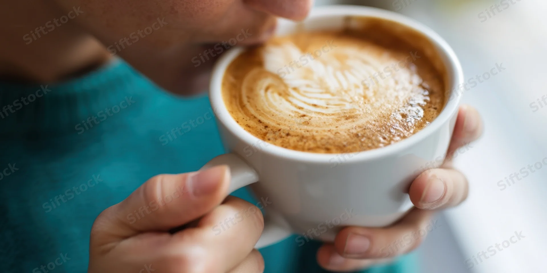 Woman sipping coffee from a white ceramic cup with latte art, hands holding cup, close-up view