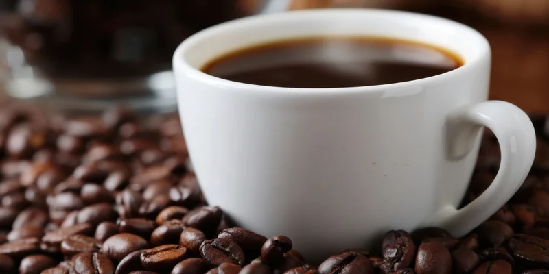 White ceramic coffee cup filled with dark coffee resting on roasted beans