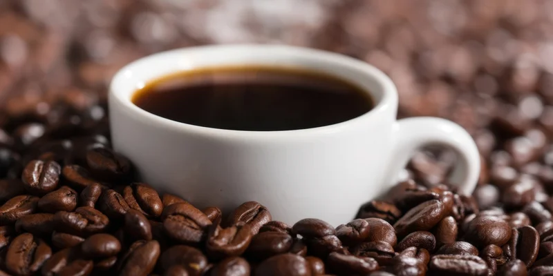 White ceramic coffee cup filled with dark coffee surrounded by roasted beans on a textured surface