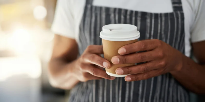Barista holding a brown disposable coffee cup with white lid, wearing striped apron over white t-shirt