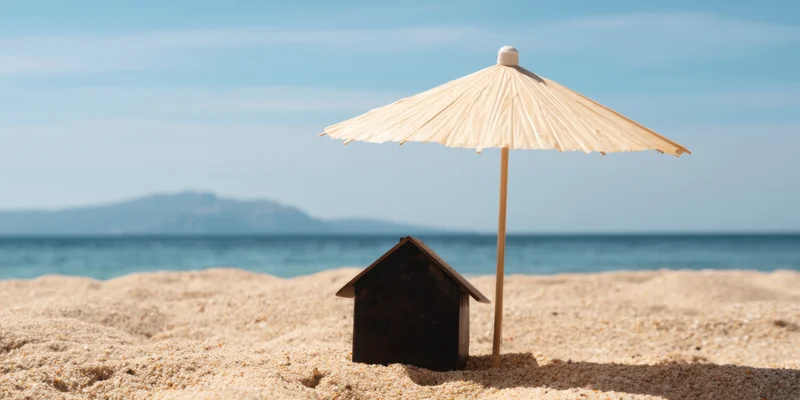 Miniature dark house with heart-shaped windows under a bamboo umbrella on sandy beach by ocean