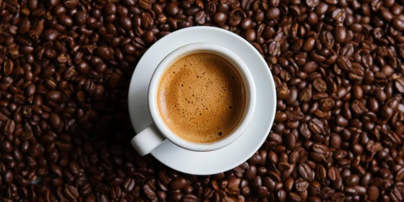 White ceramic coffee cup with crema on saucer surrounded by roasted coffee beans, top view