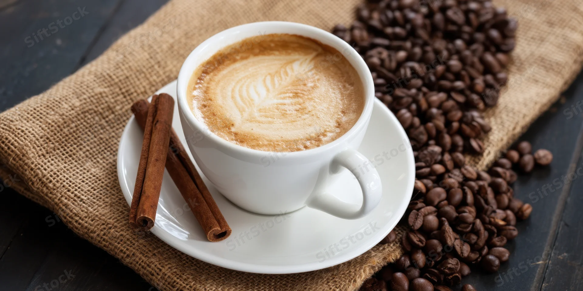 White ceramic cup of latte with leaf foam art on saucer beside cinnamon sticks and coffee beans on burlap over dark wood table