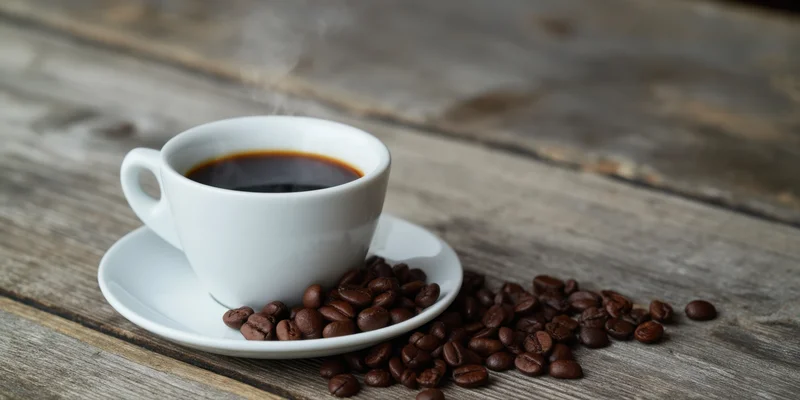 Steaming black coffee in a white ceramic cup on a wooden table with scattered coffee beans