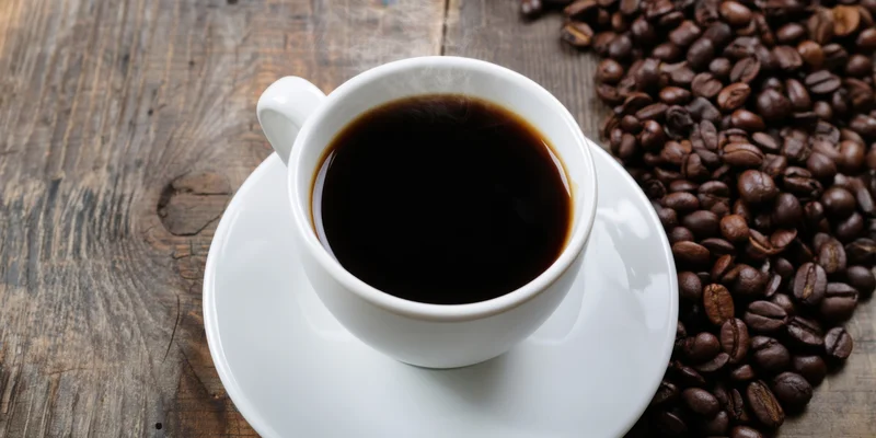 White ceramic cup of black coffee on saucer with roasted coffee beans on wooden table