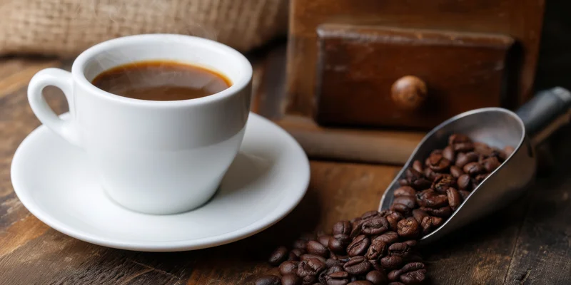 Steaming espresso cup on saucer beside coffee beans and metal scoop on wooden table