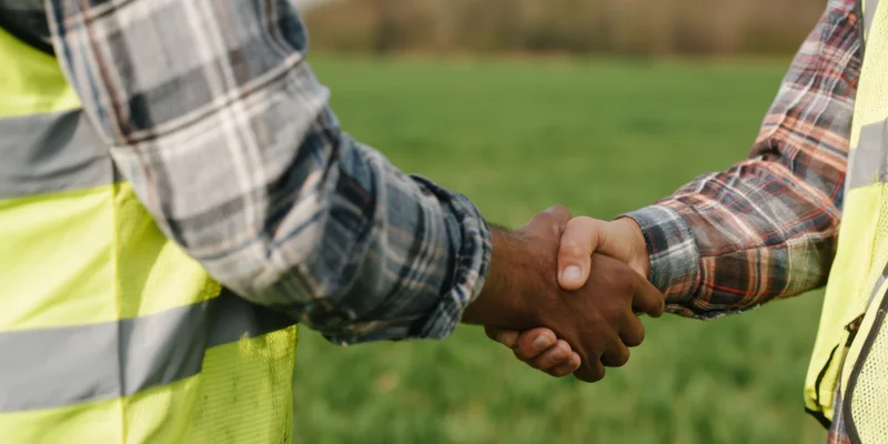 Two men in plaid shirts and safety vests shaking hands in a green field