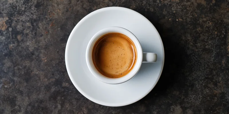 Top-down view of a white porcelain espresso cup with crema on a dark textured surface