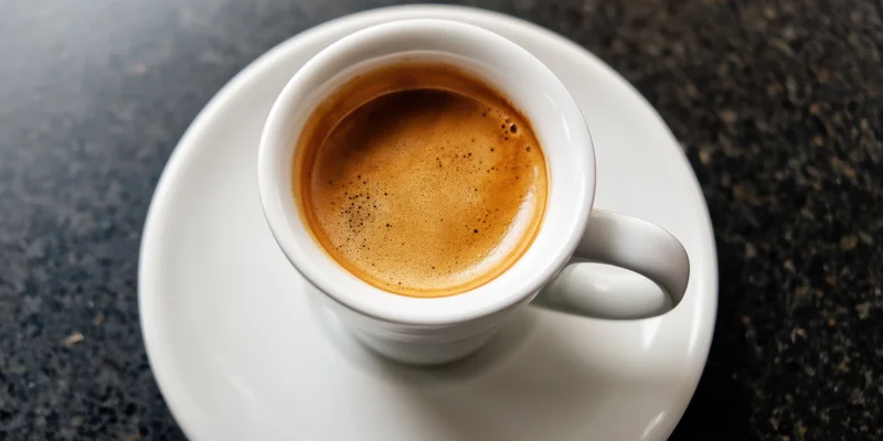 White porcelain espresso cup with crema on saucer on dark countertop, top-down view