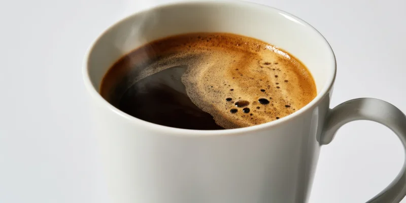 White ceramic mug filled with dark coffee and crema steam on plain white background