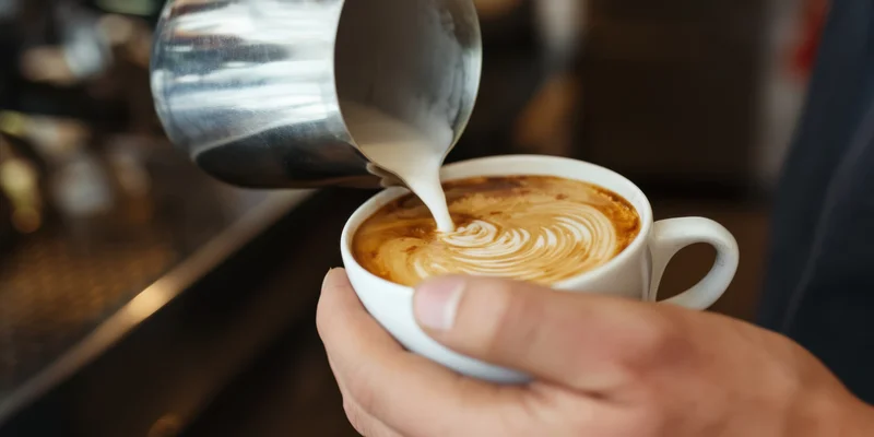 Barista pouring steamed milk into a white ceramic cup to create latte art in a coffee shop