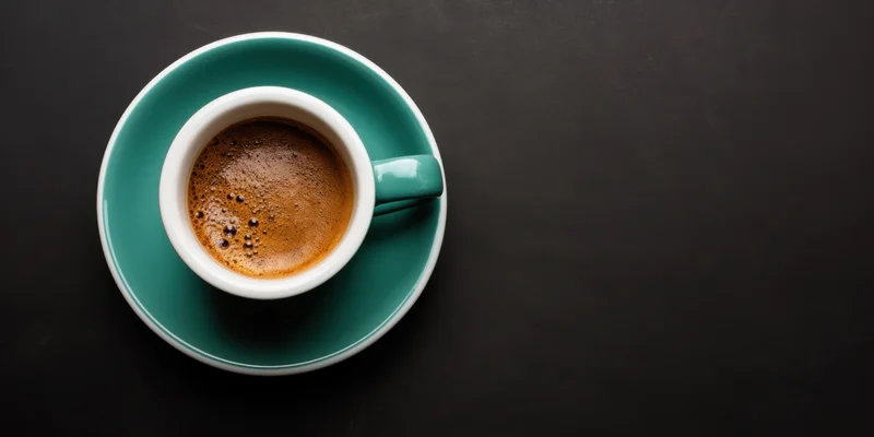 Teal ceramic coffee cup with espresso foam on matching saucer on dark surface, overhead view