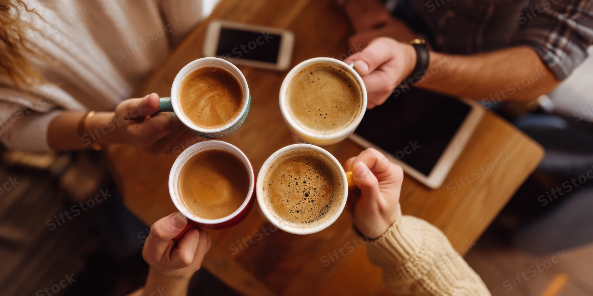 Four hands holding coffee mugs clinking together over a wooden table