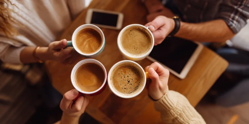 Four hands holding coffee mugs clinking together over a wooden table
