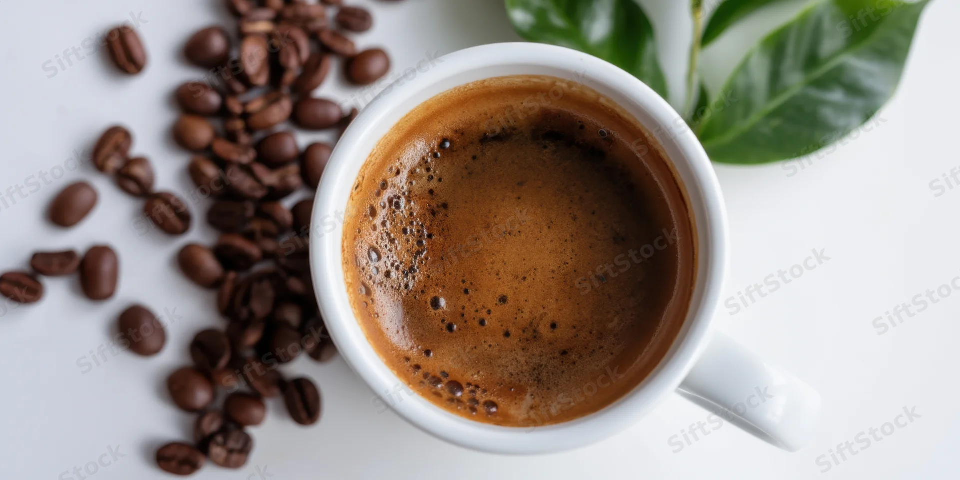 White ceramic cup filled with dark coffee and crema, surrounded by roasted beans and green leaves on a white surface