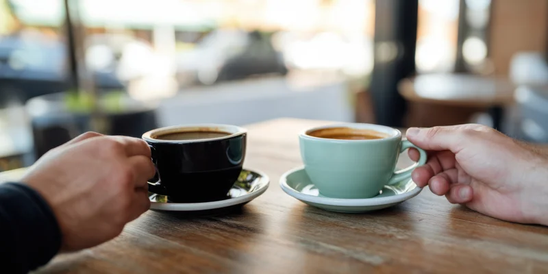 Two hands holding coffee cups on a wooden cafe table