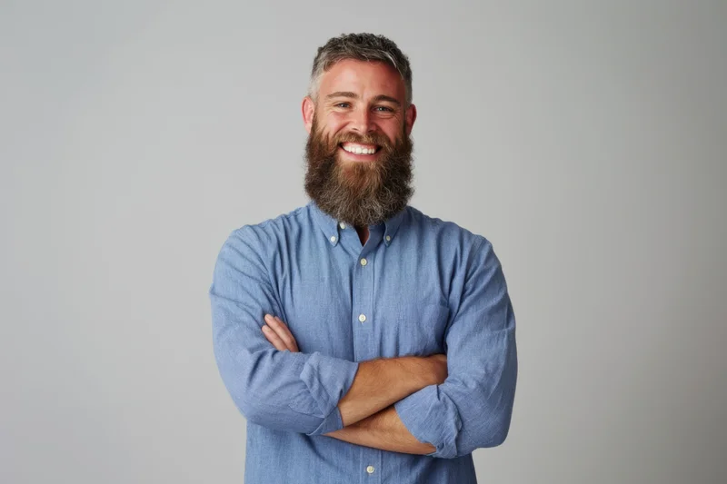 Bearded man smiling with arms crossed in blue button-down shirt against gray background