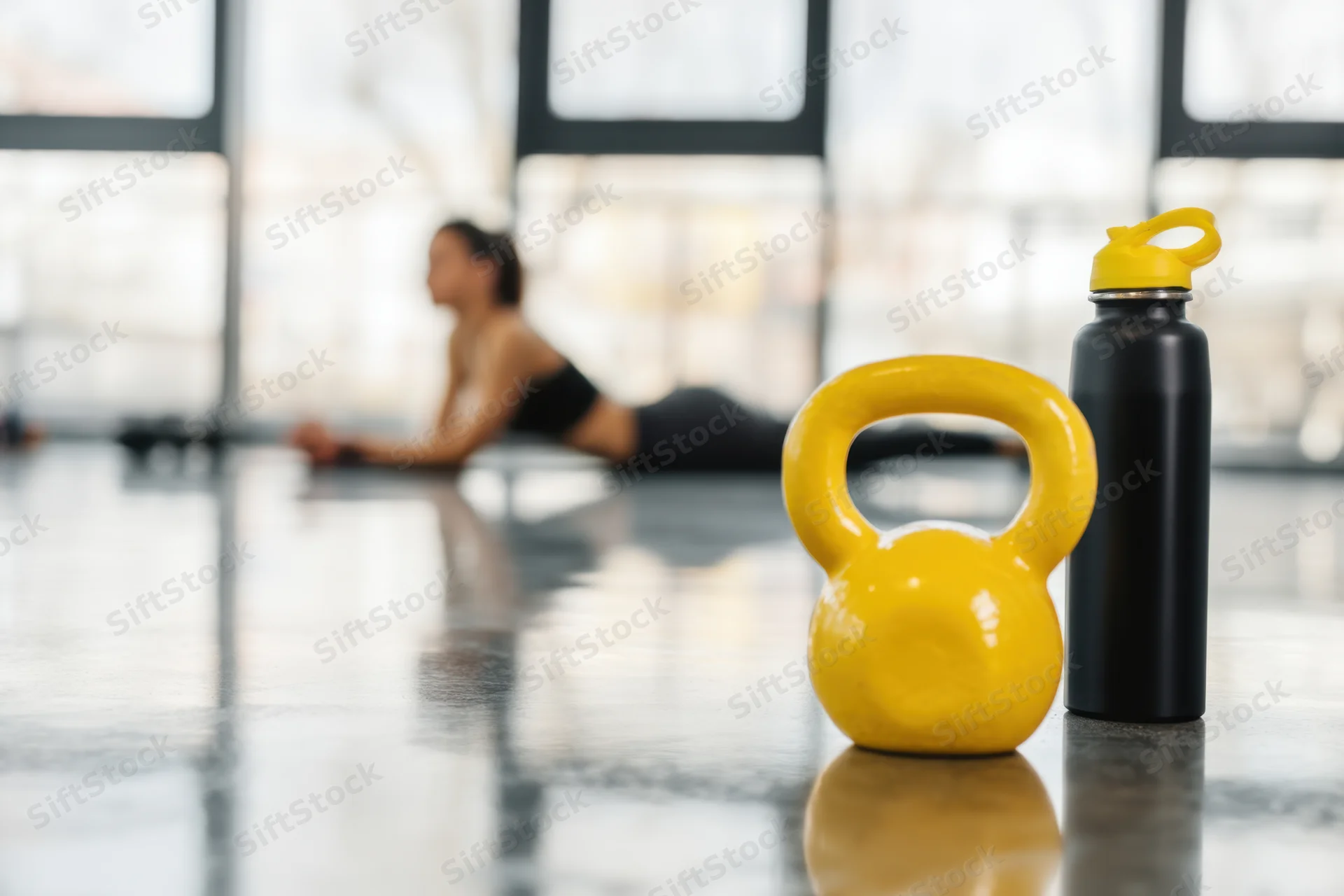 Yellow kettlebell and black water bottle on glossy gym floor with woman in plank position in background