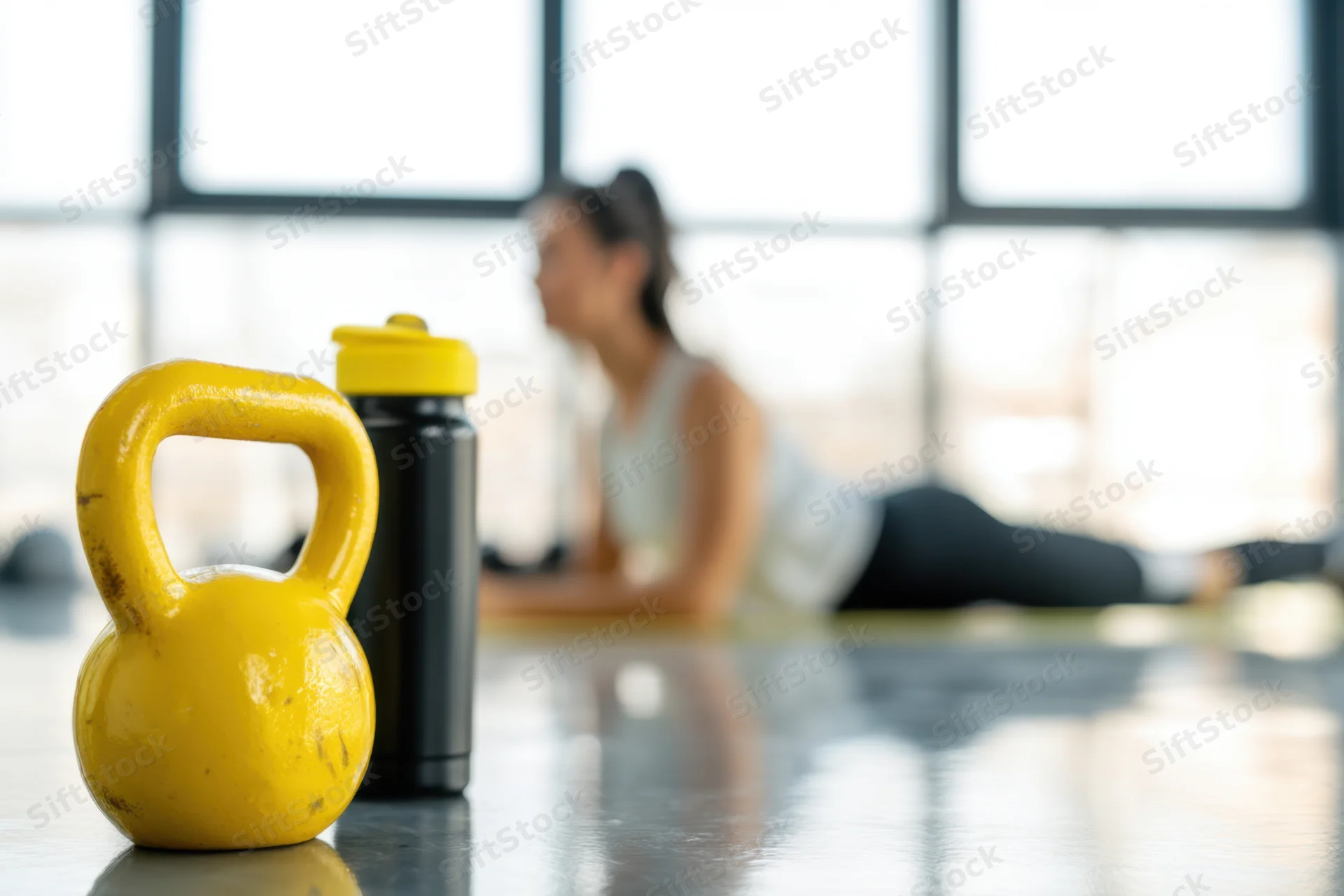 Yellow kettlebell and water bottle in foreground with woman doing plank exercise in gym background