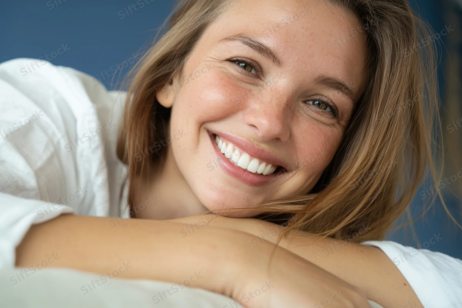 Woman smiling with light brown hair leaning on soft surface indoors