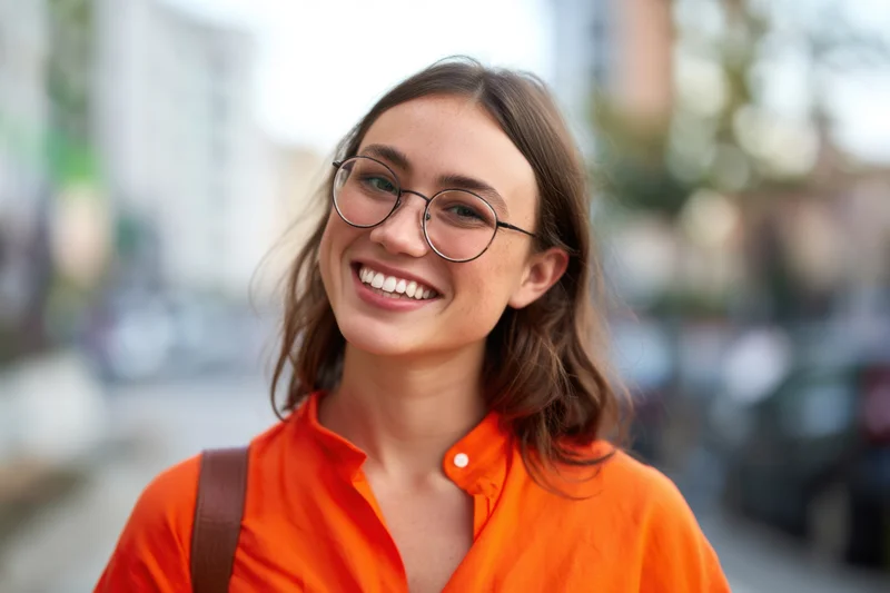 Young woman smiling with round glasses and orange shirt on city street