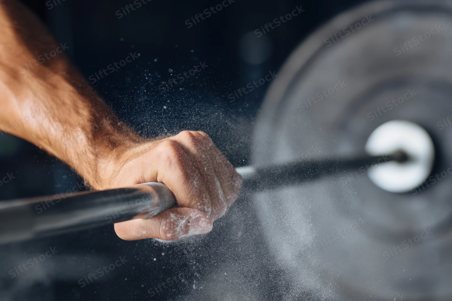 Close-up of a muscular man's hand gripping a barbell with chalk powder flying during weightlifting training in a gym