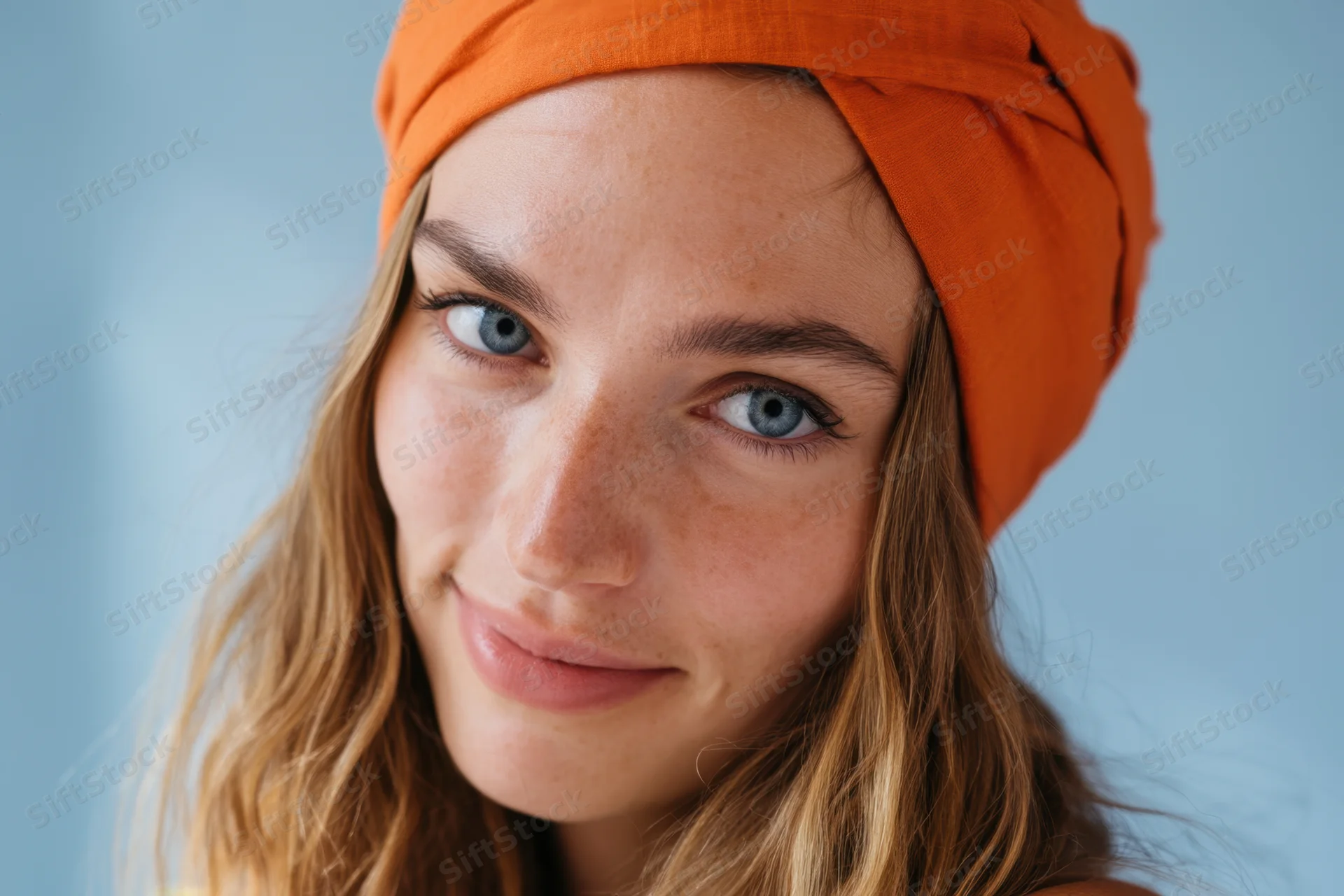 Close-up portrait of a smiling freckled woman with blue eyes and orange headband against a light blue background
