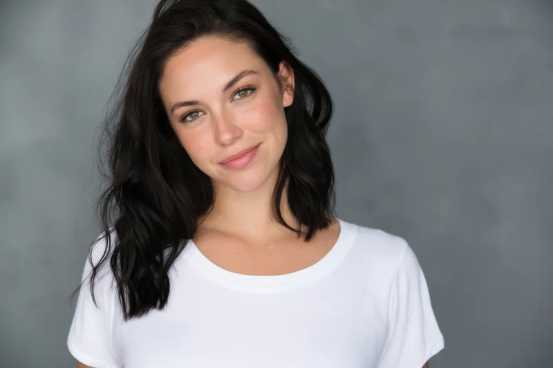 Young woman with dark wavy hair smiling softly in a white tshirt against a grey studio background
