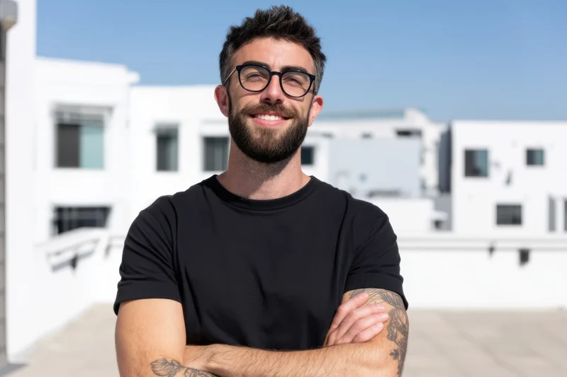 Confident man with beard and glasses smiling with arms crossed on rooftop against white buildings and blue sky
