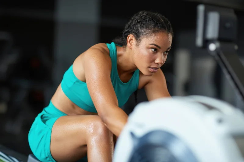 Woman exercising on rowing machine in gym, wearing teal sports bra and shorts, focused expression