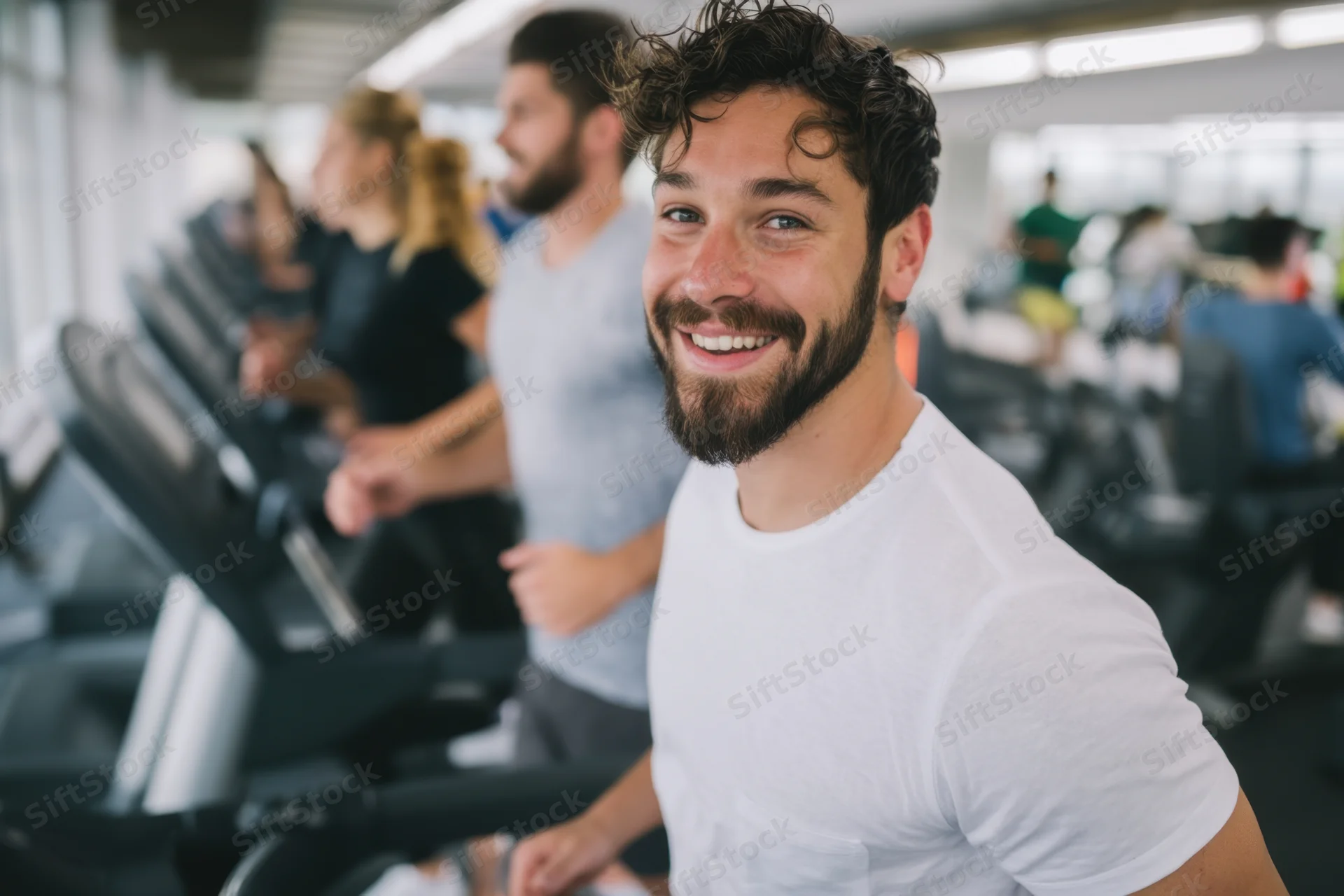 Man smiling while running on treadmill in bright gym with others exercising in background