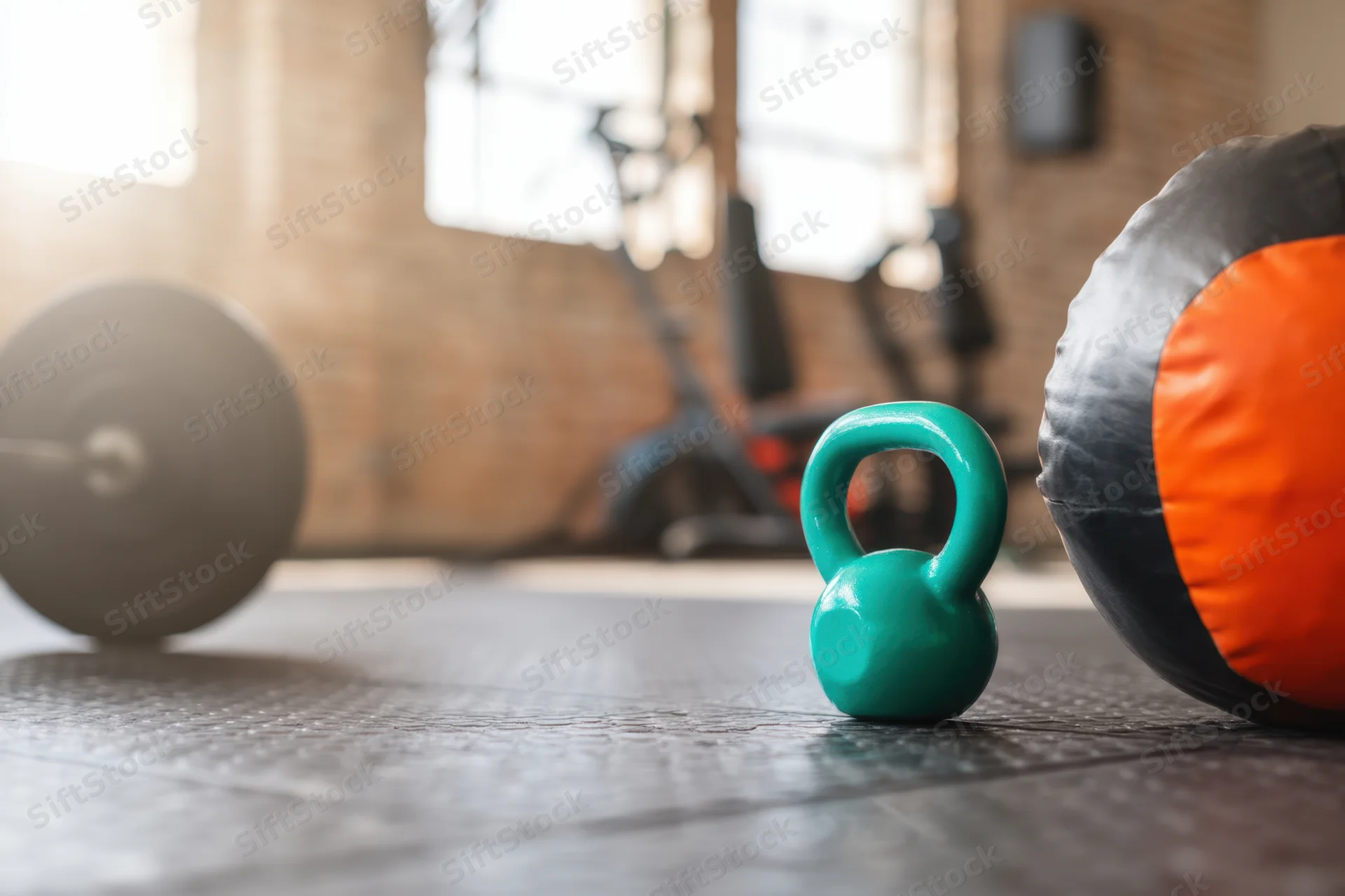 Teal kettlebell and orange medicine ball on textured gym floor with barbell plate and exercise equipment in background