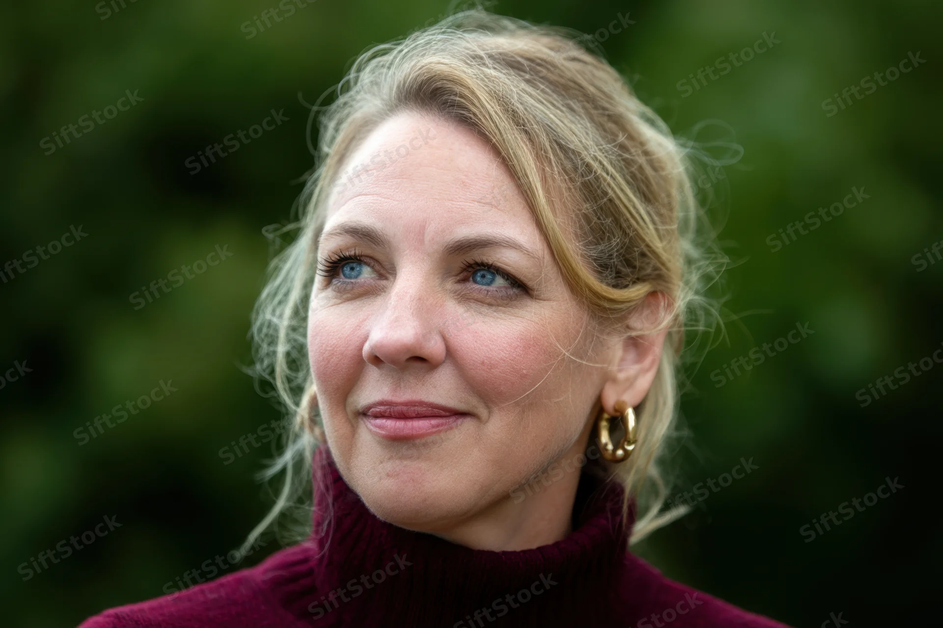 Woman with blonde hair in a maroon turtleneck smiling softly outdoors