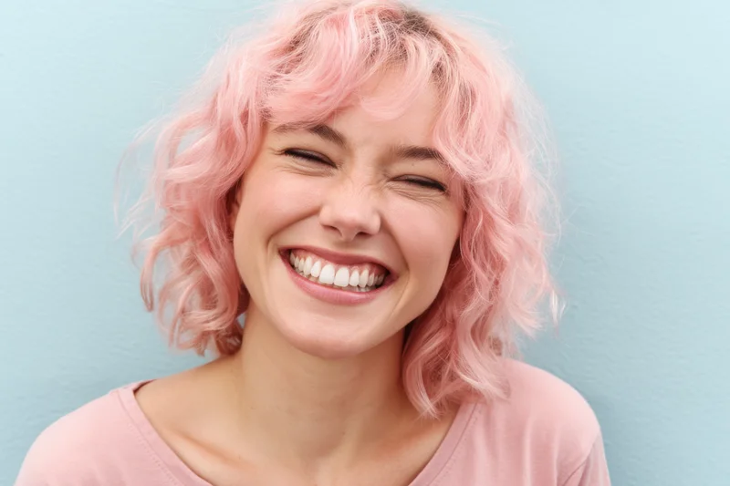 Woman with pastel pink curly hair smiling joyfully in closeup against blue background