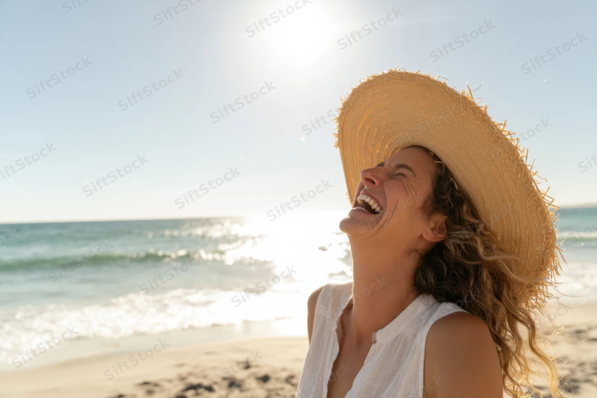 Woman laughing on sunlit beach wearing straw hat and white tunic