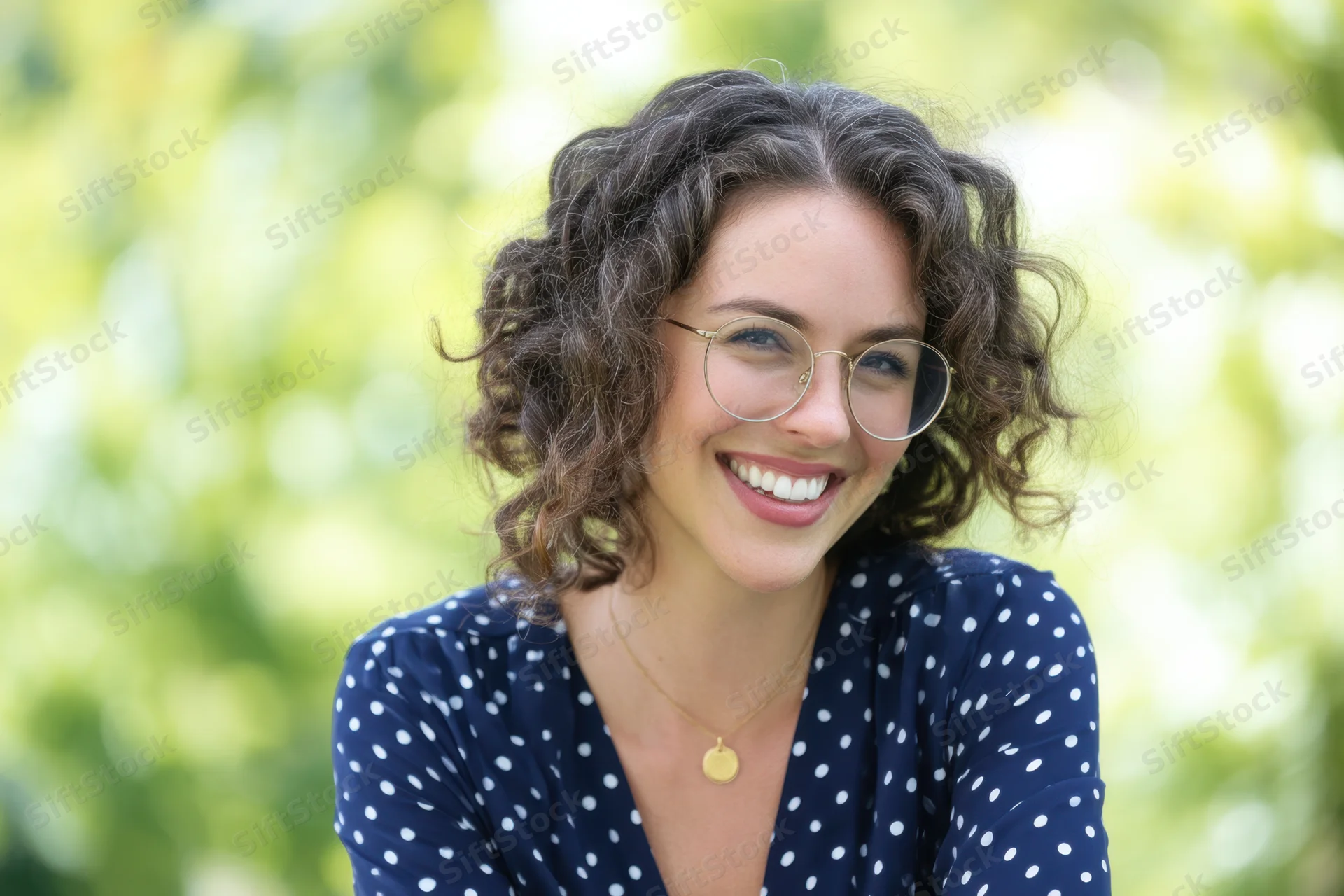 Smiling woman with round glasses and curly hair outdoors in a green setting