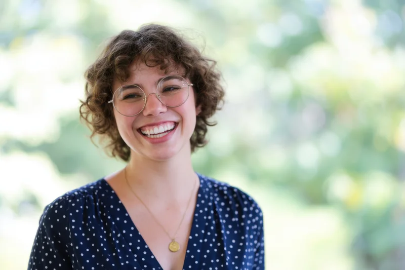 Smiling woman with round glasses and curly hair outdoors in a polka dot blouse
