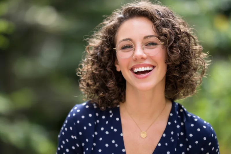 Woman with curly hair and round glasses smiling joyfully outdoors