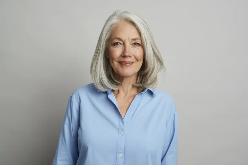 Senior woman with shoulder-length silver hair wearing a light blue button-up shirt, smiling calmly in a studio portrait setting