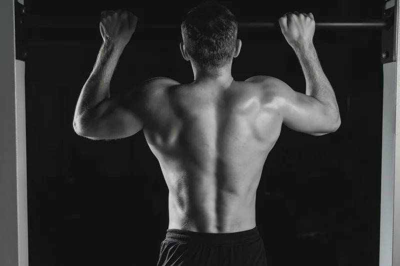 Muscular man hanging from pull-up bar in dark gym showing back and arms