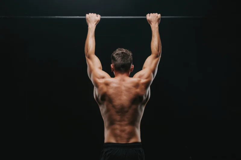 Muscular man hanging from pull-up bar in dark gym showing back muscles