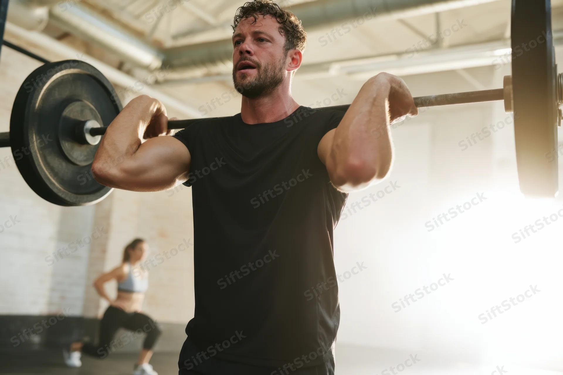 Man lifting barbell in gym with woman exercising in background