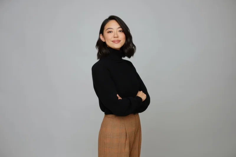 Asian woman standing with arms crossed in black turtleneck and plaid skirt against gray background