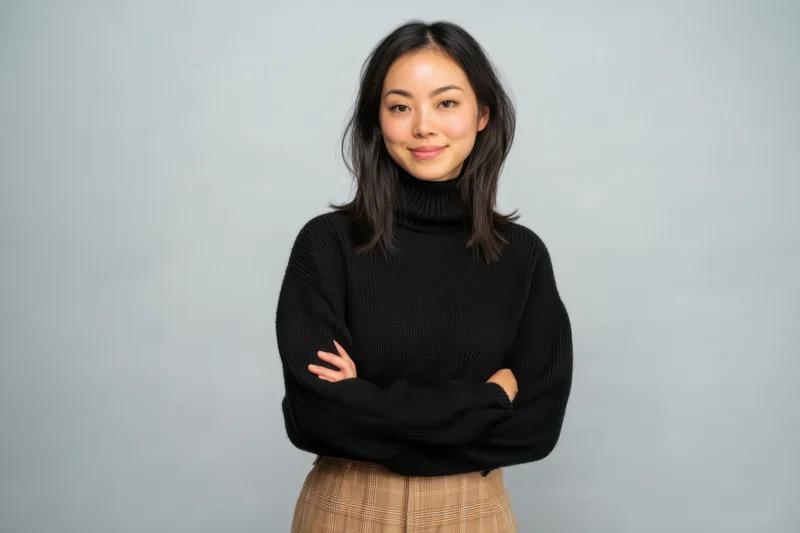 Asian woman standing with arms crossed in black turtleneck sweater and plaid skirt against gray studio background