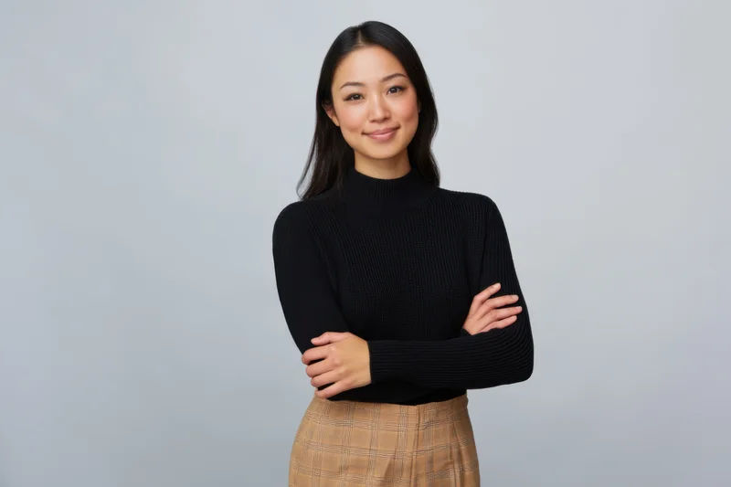 Asian woman standing with arms crossed in studio wearing black turtleneck sweater and plaid skirt